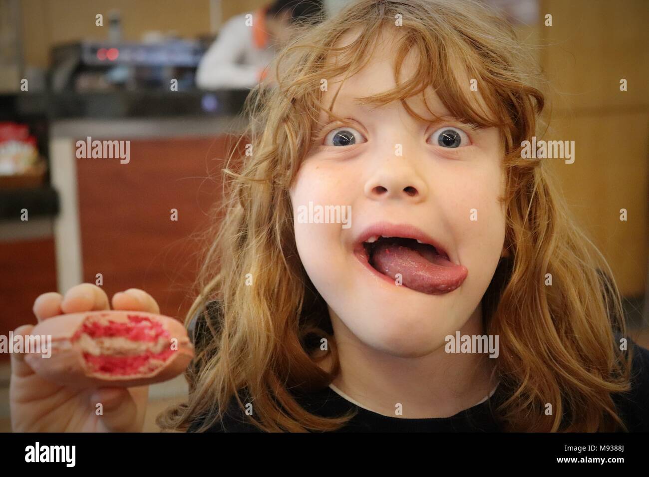 Una ragazza con lo zenzero Ricci i capelli e gli occhi blu bastoni la sua lingua fuori come lei mangia un over - dimensioni Macaron rosa Foto Stock