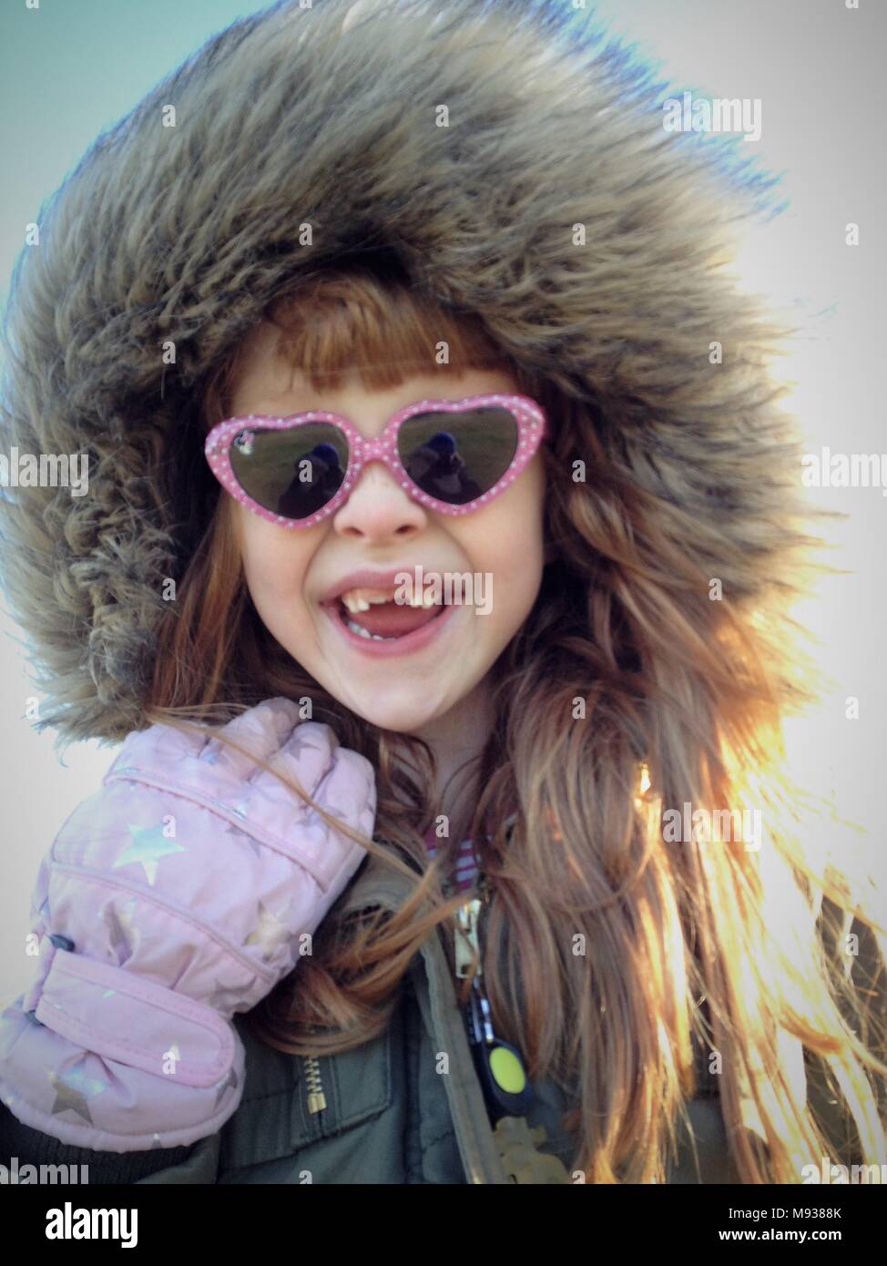 Una ragazza con lunghi capelli di zenzero e Gap denti indossa rosa a forma di cuore occhiali, guanti rosa e e una cappa di pelliccia Foto Stock