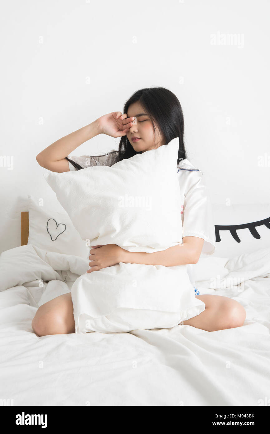 Assonnato giovane ragazza asiatica chiuse gli occhi e seduta sul letto con cuscino in camera bianca. Foto Stock