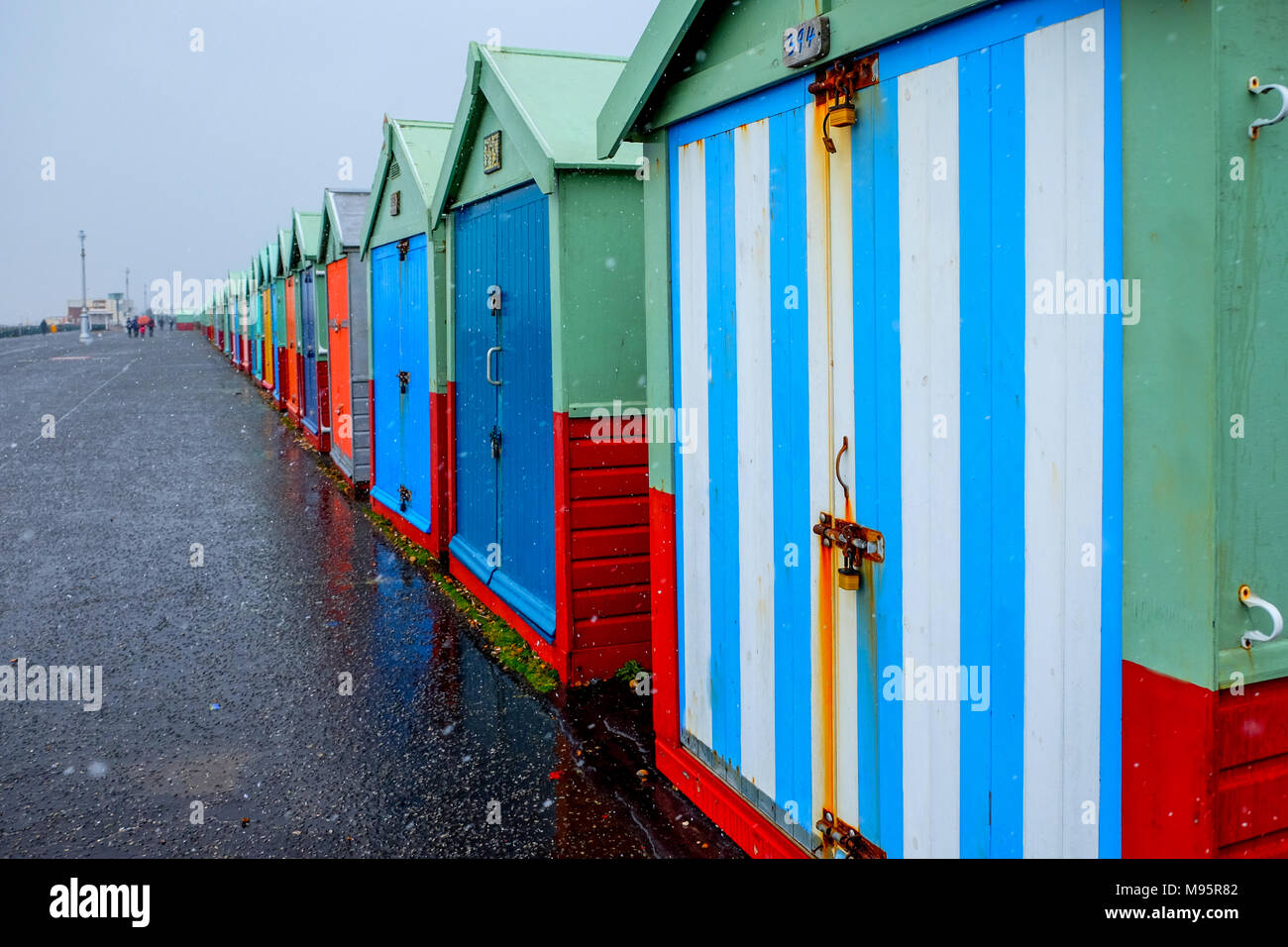 Brighton Seafront quaranta beach capanne, le capanne sono variopinte porte in una linea retta su un calcestruzzo promenade il cielo grigio è la spiaggia più vicina h Foto Stock