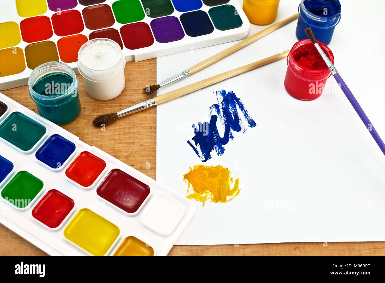 Due scatole di vernice, tre spazzole, vasi con gouache, un foglio di carta con un'immagine su una tavola di legno Foto Stock