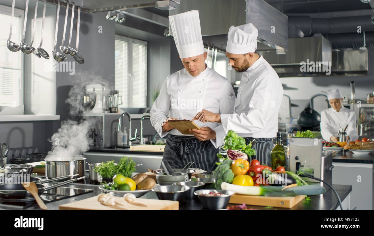 Due famosi chef discutere i loro blog video mentre si utilizzano computer Tablet. Essi lavorano su un grande ristorante in Acciaio Inox cucina professionale. Foto Stock