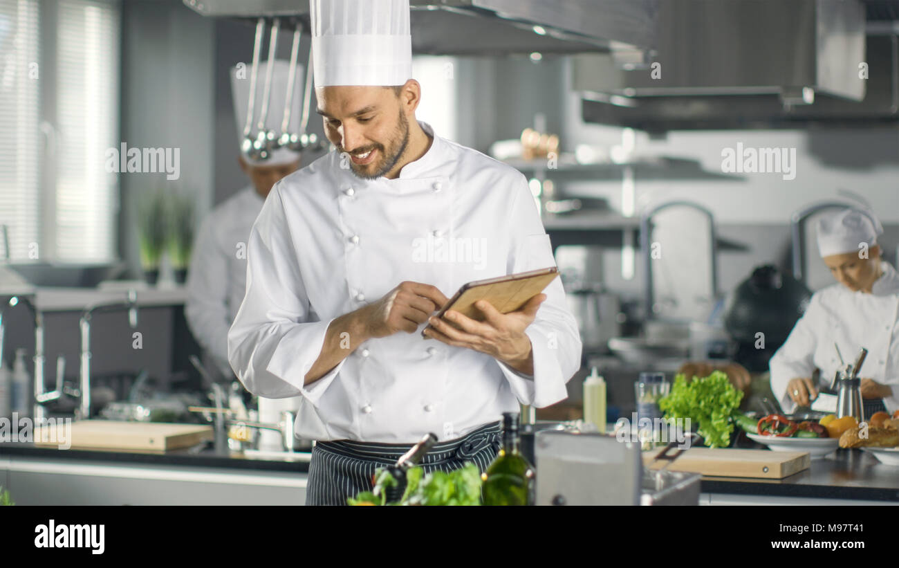 Famoso Chef utilizza computer Tablet per ricette mentre si lavora in una cucina moderna. Il suo aiuto opera in background. Foto Stock