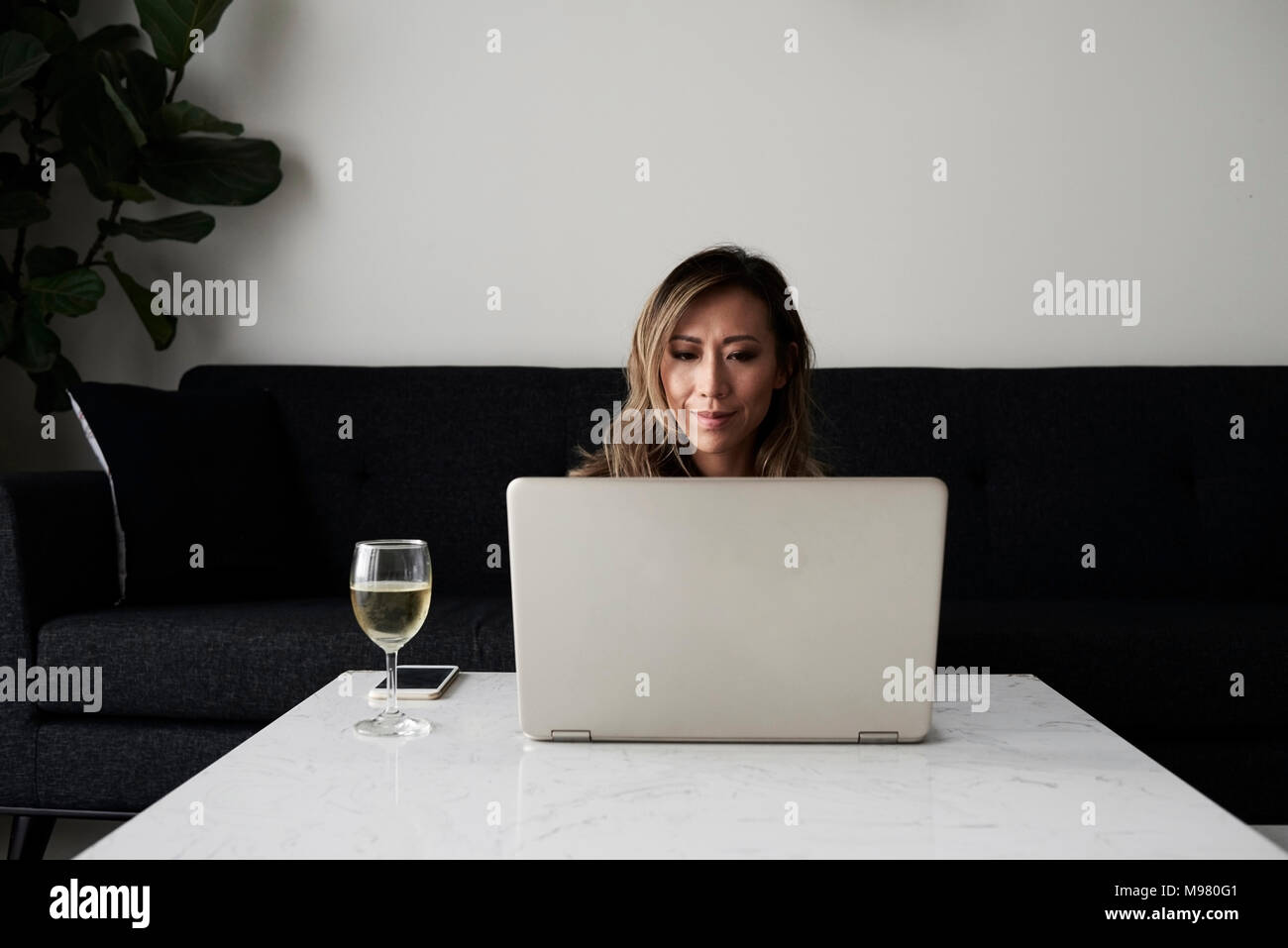 Bella donna asiatica avente un bicchiere di vino mentre si lavora con il portatile a casa. Foto Stock