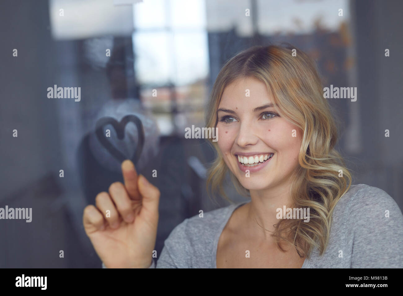 Ritratto di felice donna bionda cuore di disegno sul vetro di finestra Foto Stock