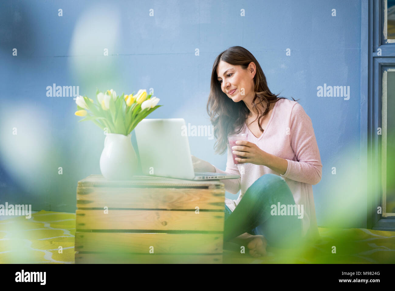Donna seduta sul pavimento con bicchiere di succo di frutta con laptop Foto Stock