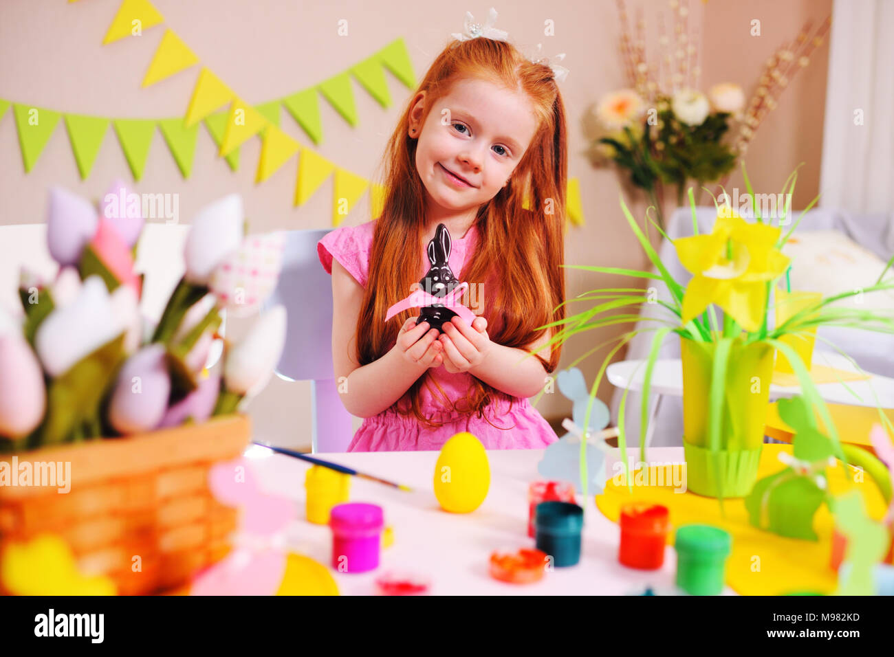 Baby piccola ragazza con i capelli rossi con cioccolato coniglietto di pasqua in mani su di uno sfondo di decorazioni di Pasqua Foto Stock