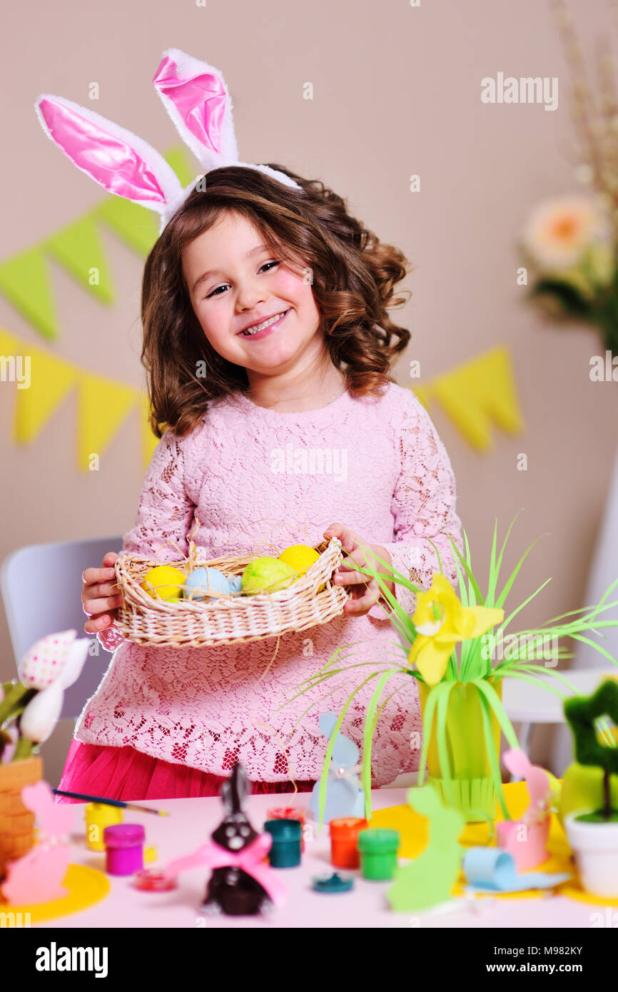 Bambina con orecchie di coniglio in possesso di un cesto di Pasqua con dipinto di uova colorate e sorridente sullo sfondo delle decorazioni di Pasqua Foto Stock