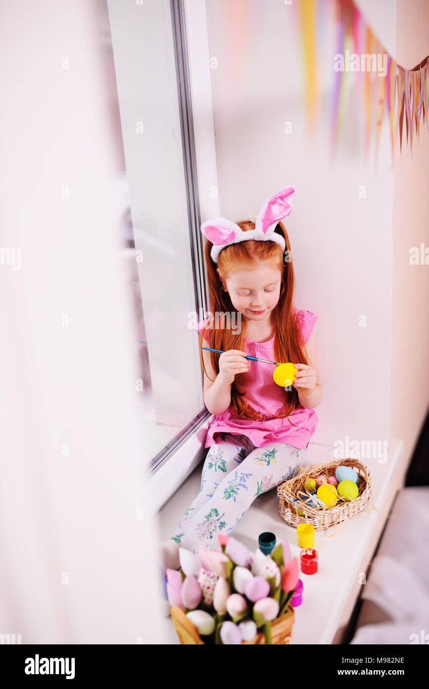 Una piccola bella ragazza con i capelli rossi con orecchie di coniglio sul suo capo è la pittura delle uova per un cesto di Pasqua sul davanzale contro la finestra e la Pasqua d Foto Stock