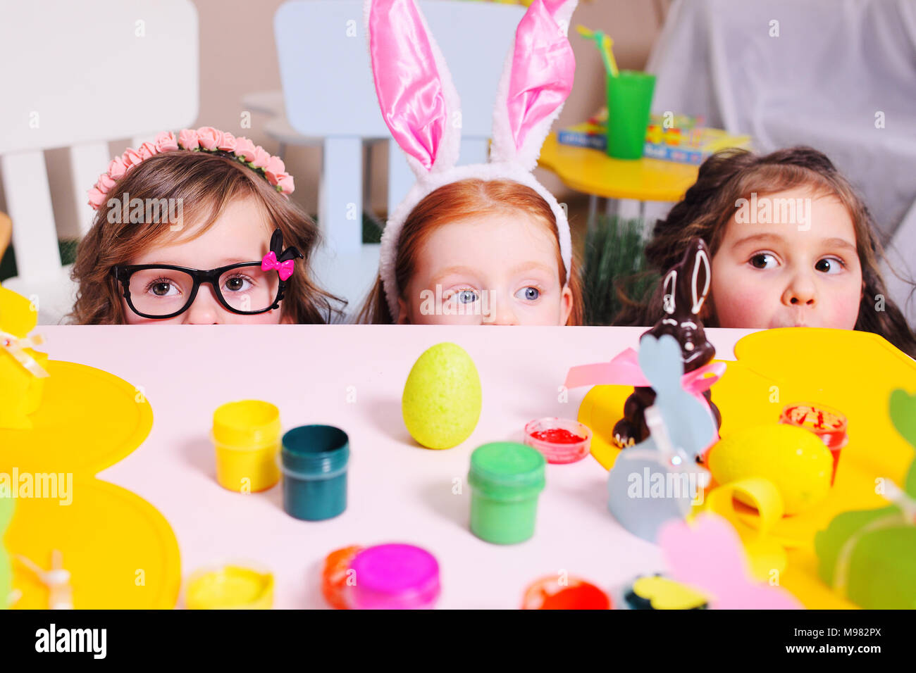 I bambini nel coniglio le orecchie peek fuori da sotto il tavolo sullo sfondo della Pasqua decor. Preparazione per la celebrazione della Pasqua. Caccia Foto Stock