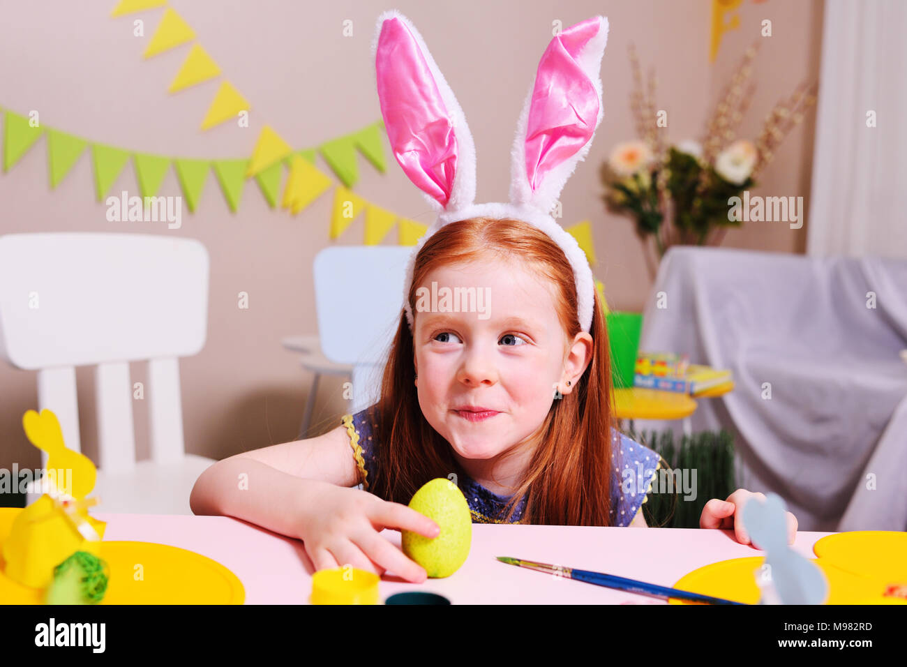 Allegro bambino attivo la ragazza con i capelli rossi e orecchie di coniglio dipinge un uovo con vernice gialla su uno sfondo di pasqua decor e sorrisi.Le tradizioni pasquali Foto Stock