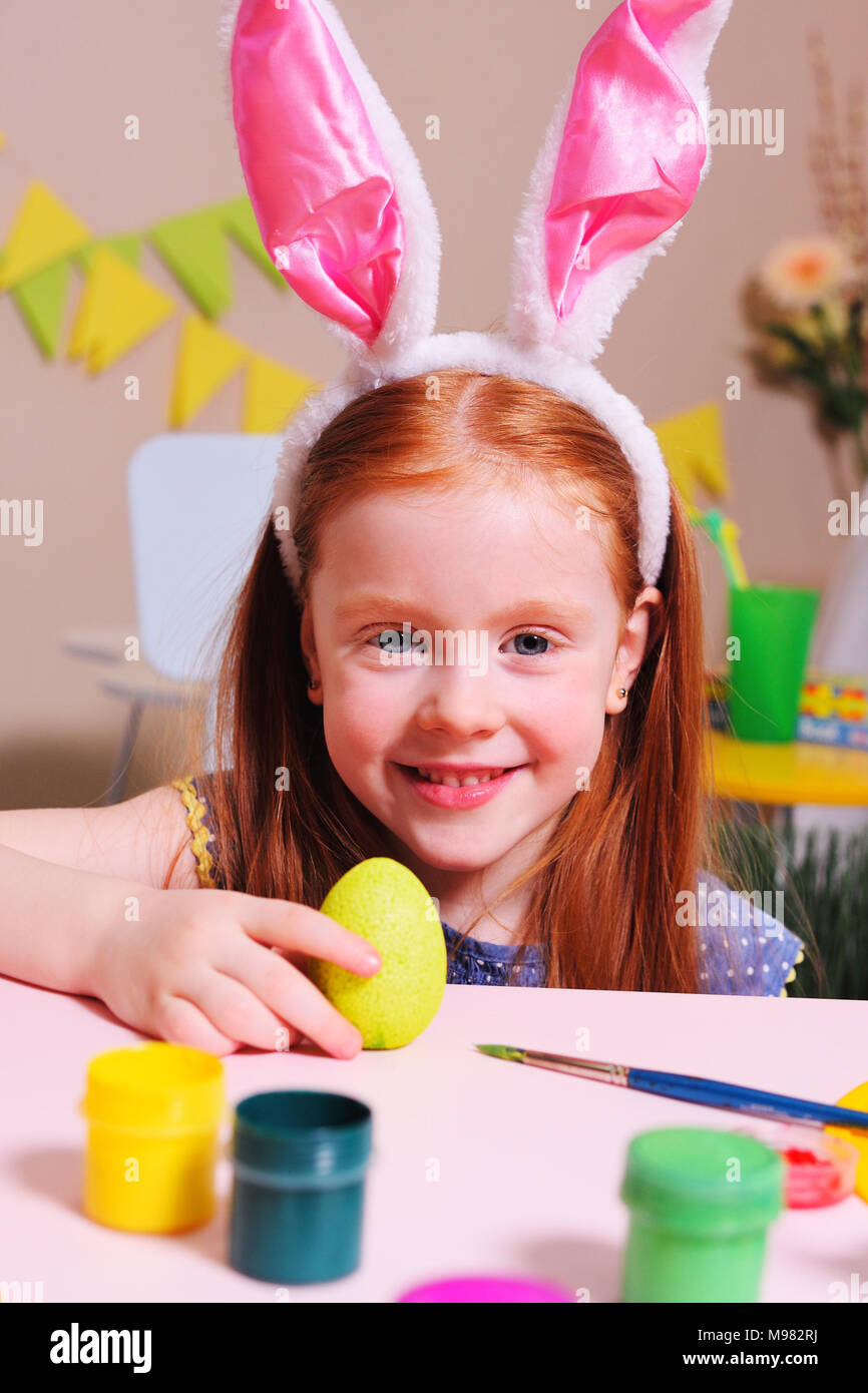 Allegro bambino attivo la ragazza con i capelli rossi e orecchie di coniglio dipinge un uovo con vernice gialla su uno sfondo di pasqua decor e sorrisi.Le tradizioni pasquali Foto Stock