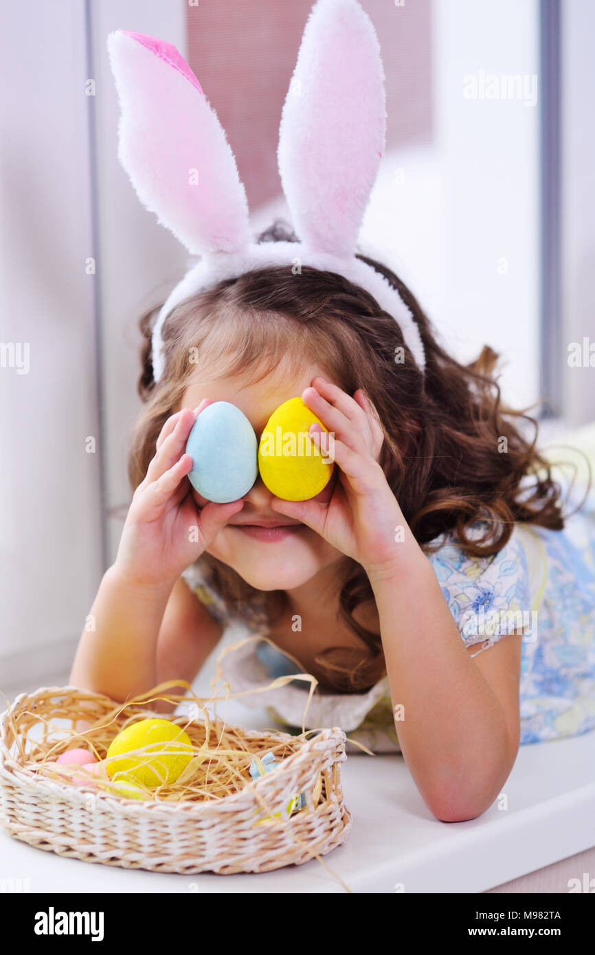 Bambina con orecchie di coniglio con uova dipinte in mani sorridente sullo sfondo del cesto di pasqua seduto vicino a finestra Foto Stock