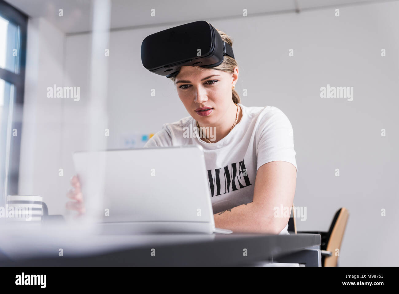 Giovane donna con laptop e occhiali VR lavorare alla scrivania in ufficio Foto Stock