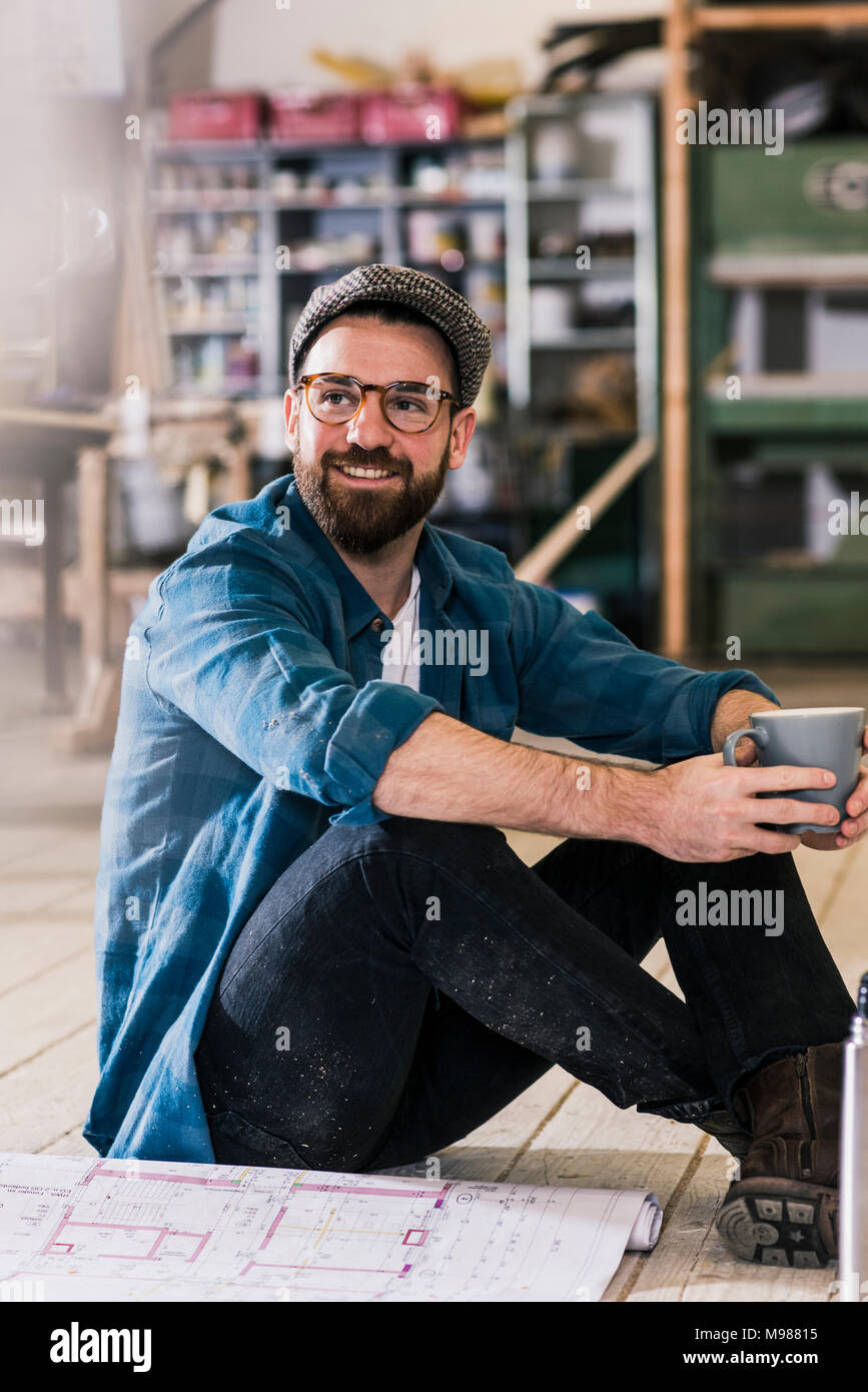 Uomo sorridente con tazza di caffè e piano di costruzione seduta sul pavimento Foto Stock