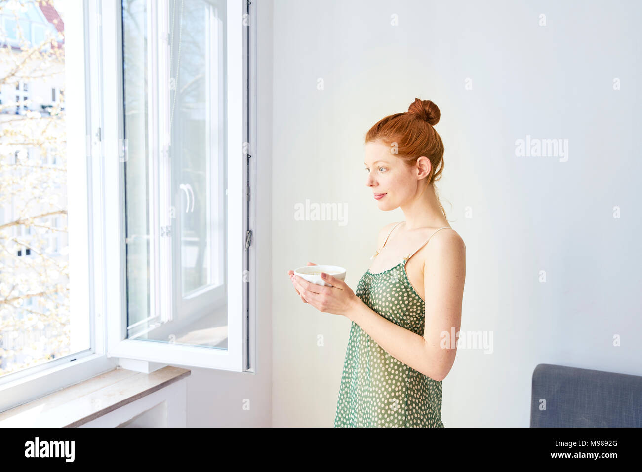 Ritratto di donna pensieroso con tazza di caffè bianco in piedi vicino alla finestra aperta Foto Stock
