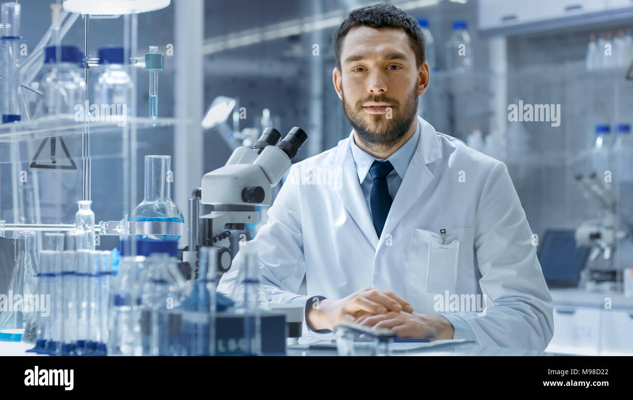 Giovane Maschio Research guarda nella telecamera e sorrisi. Egli è seduta in una fascia alta laboratorio moderno con bicchieri, vetreria, microscopio e monitor di lavoro Foto Stock