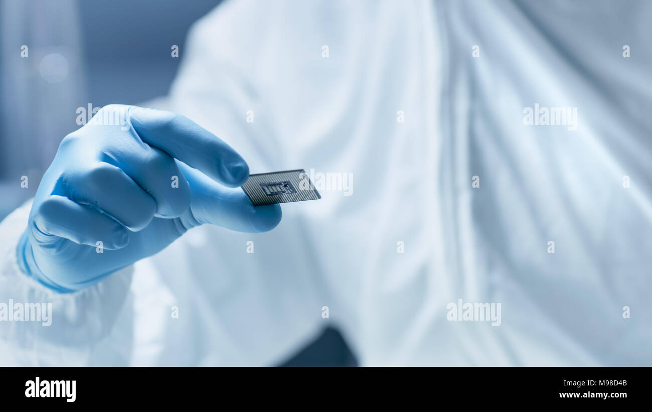 In ultra moderne Electronic Manufacturing Factory Design Engineer in tuta sterile detiene il microchip con guanti e lo esamina. Foto Stock