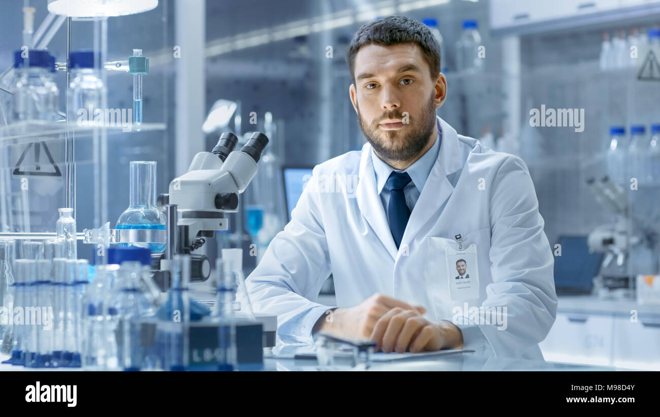 Giovane Maschio Research Scientist colloqui nella fotocamera. Egli è seduta in una fascia alta laboratorio moderno con bicchieri, vetreria, microscopio e monitor di lavoro Foto Stock