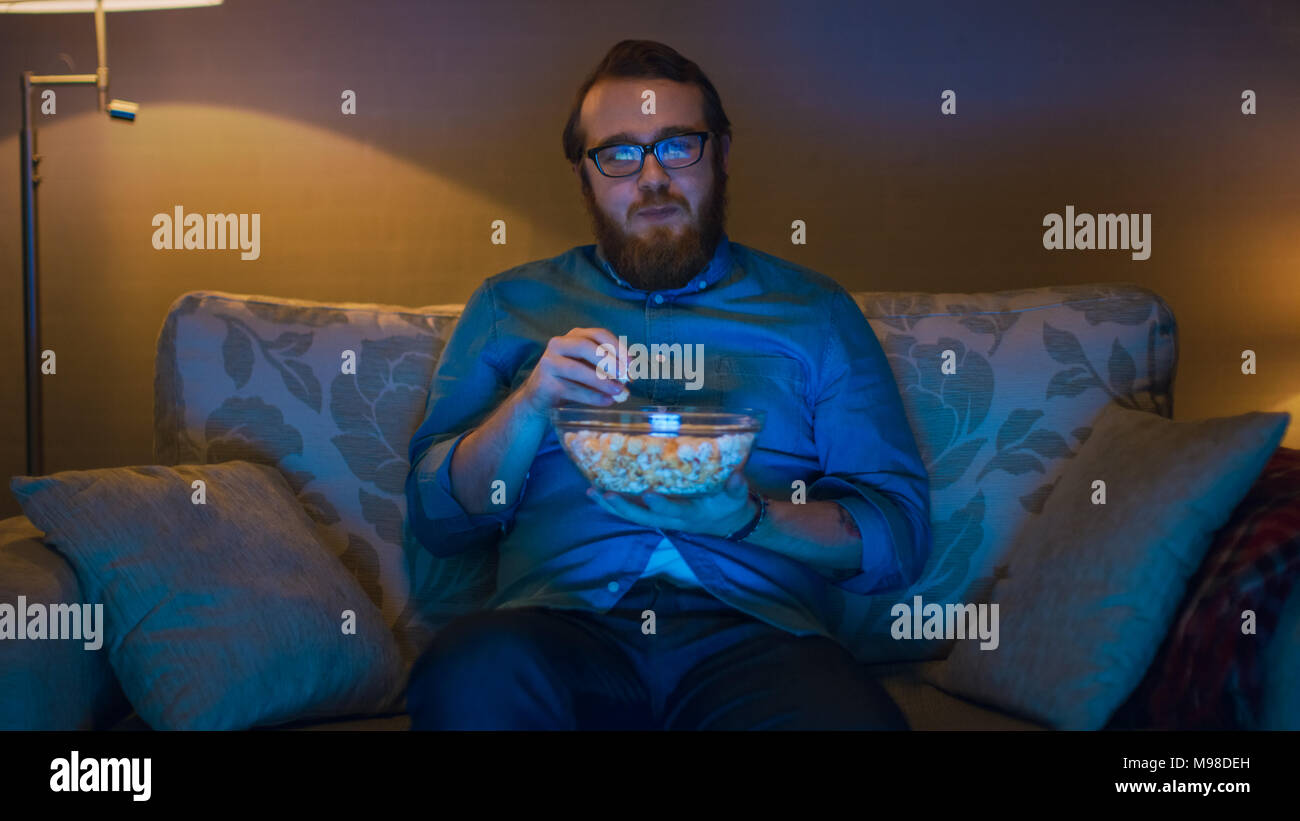 Ritratto di un uomo seduto su un divano nel salotto di casa sua, mangiare popcorn e guardare la TV. Lampade da terra sono accesi. Foto Stock