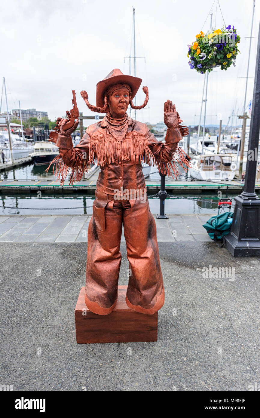 Artista di strada vestito come una statua di cowboy in costume marrone e cappello, in posa con pistole vicino a un porticciolo con yacht e barche. Foto Stock
