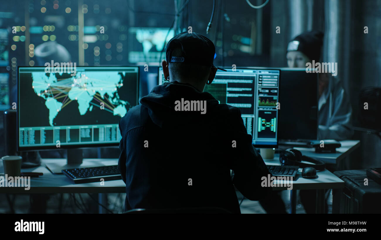 Vista posteriore di hacker adolescenti che lavorano in Computer e infettare con virus server dei dati delle infrastrutture del governo. Il suo nascondiglio è scuro con monitor Foto Stock