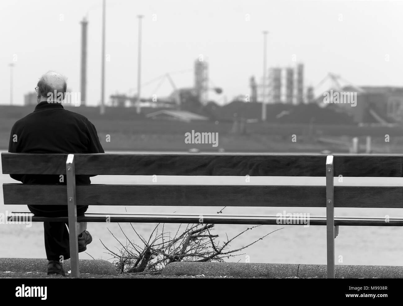 Uomo si siede solitaria su un banco di sinistra con industria pesante in background Foto Stock