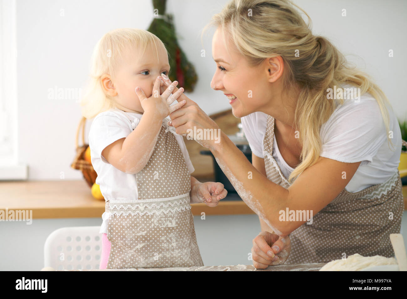 Bambina e la sua bionda in mom grembiuli rossi a giocare e ridere mentre impastare la pasta in cucina. In casa la pasta per pane e pizza o cuocere i biscotti in forno Foto Stock