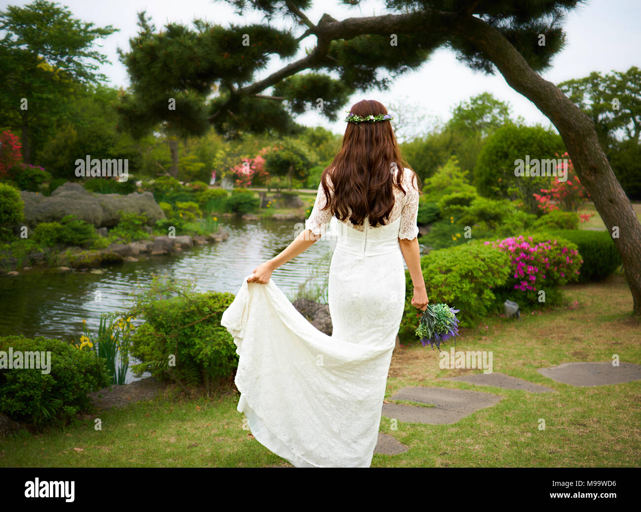 Bella donna asiatica in un bianco abito da sposa indossa una corona di fiori in possesso di un mazzo di fiori viola e mostrando il suo ritorno in un giardino con laghetto e pino Foto Stock