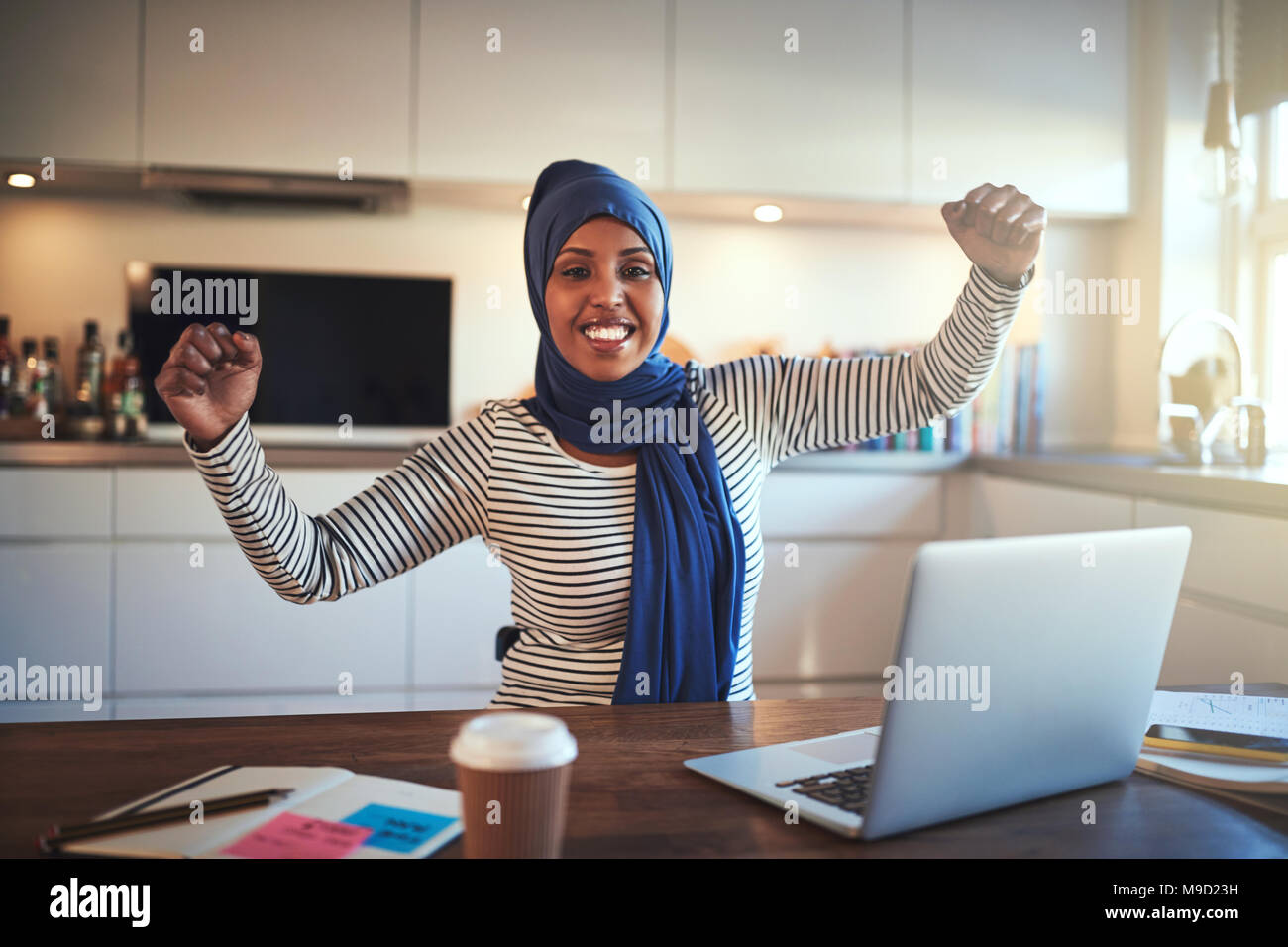 Estasiato giovani musulmani imprenditore femmina indossando un hijab alzando le braccia in successo mentre si lavora su un computer portatile nella sua cucina Foto Stock