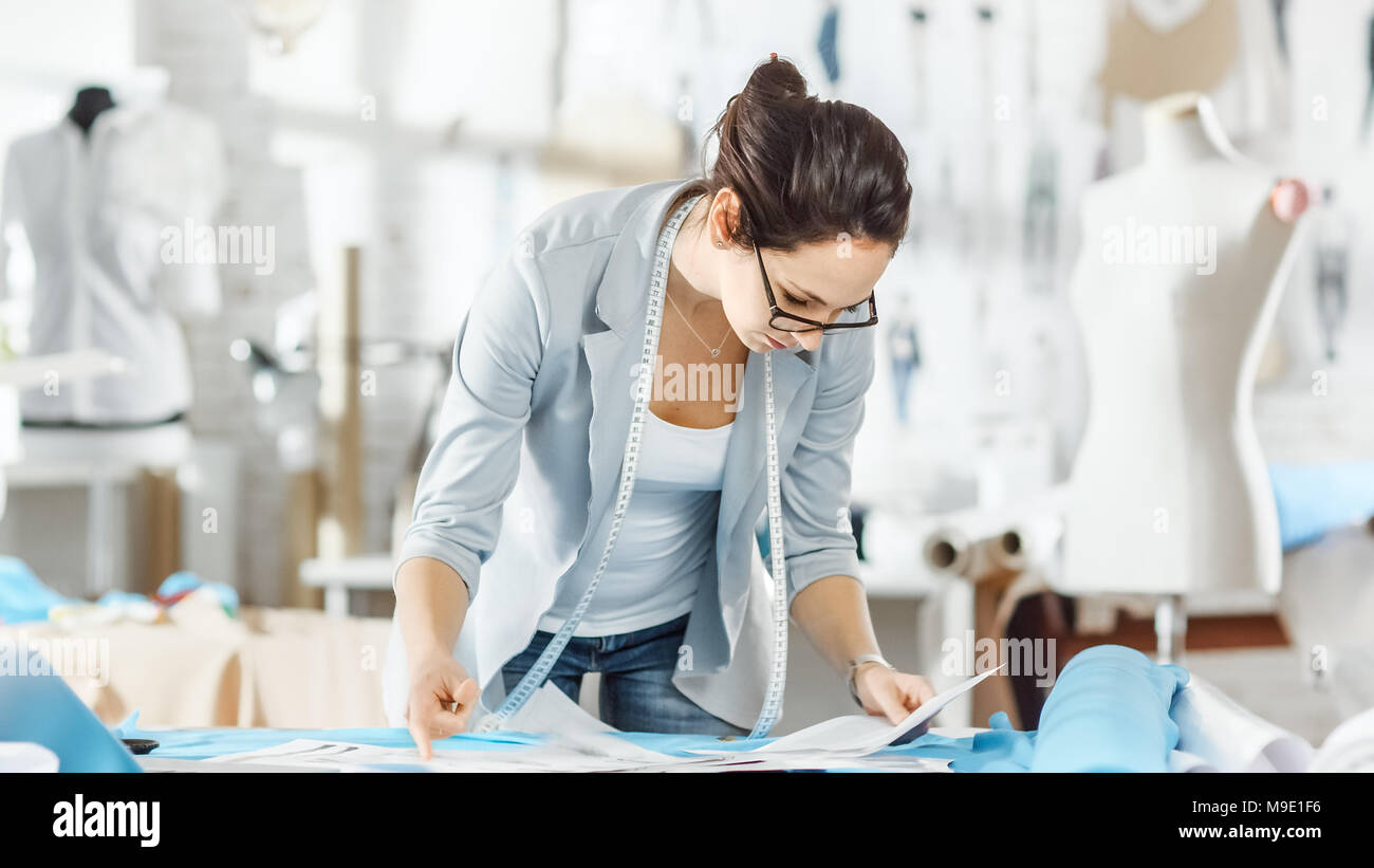 Bella la moda, progettista sceglie tra schizzi utilizza il suo computer portatile. Tessuti e articoli di cucitura che stabilisce intorno a lei. Studio è soleggiato. Foto Stock