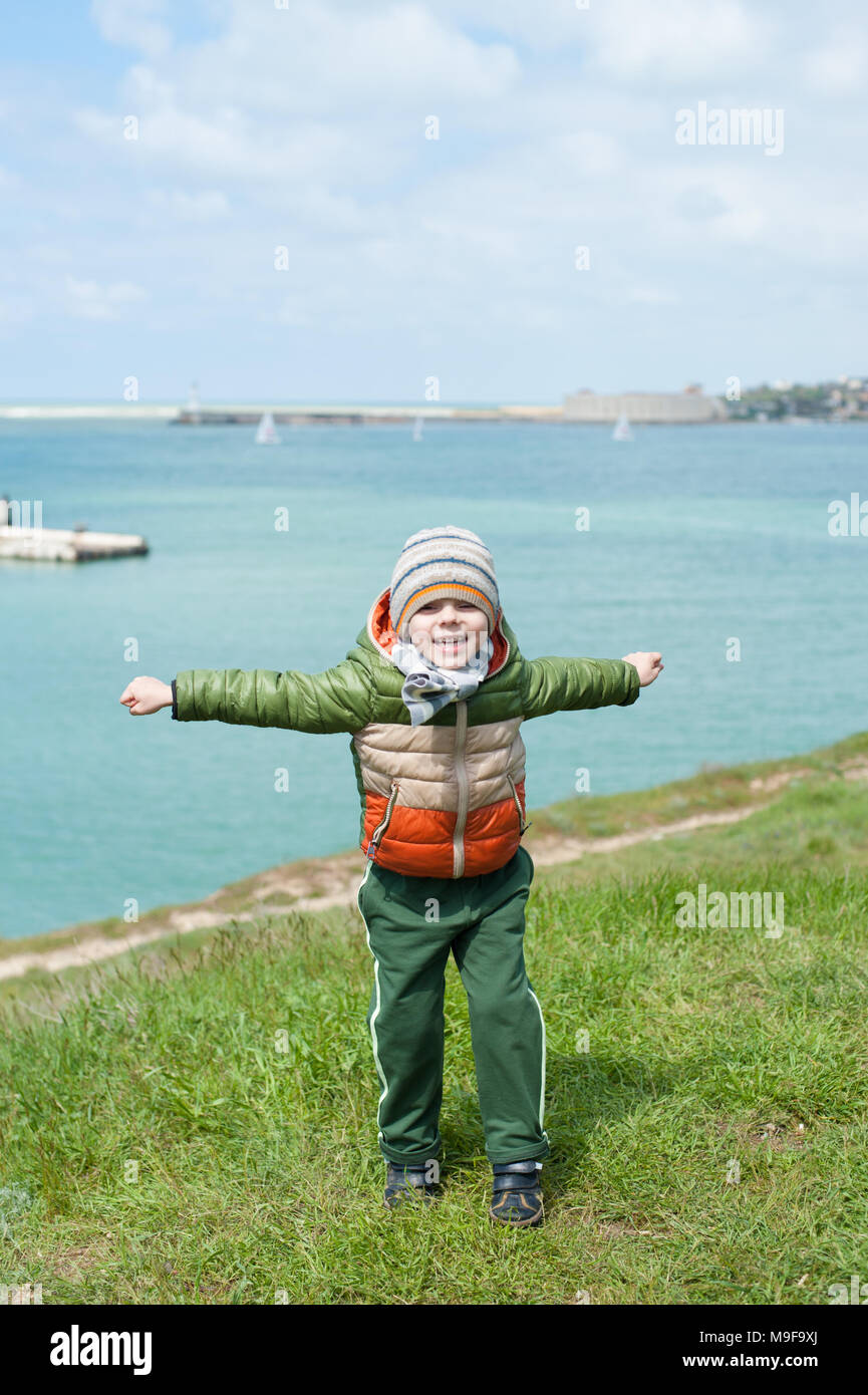 Carino piccolo ragazzo vestito in giacca calda sciarpa e hat salta sul verde pendio sullo sfondo del porto di mare con barche in Cold spring Foto Stock