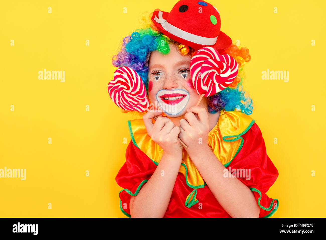 Closeup ritratto di capretto piccolo indossare tuta da clown, tenendo lollipop. Studio shot, isolato su sfondo giallo Foto Stock