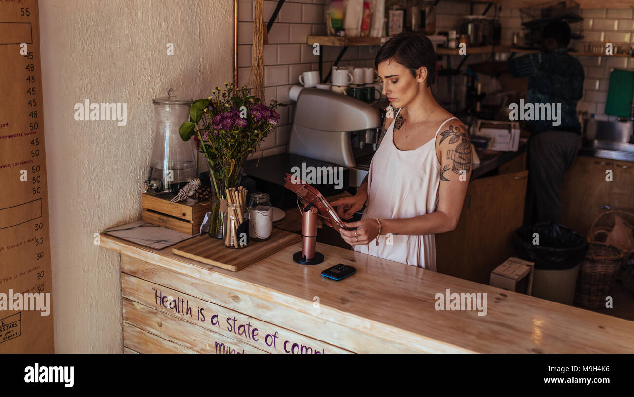 Donna in piedi presso il contatore di fatturazione del suo coffee shop. Coffee shop proprietario il funzionamento della macchina di fatturazione mentre un lavoratore si prepara il caffè in backgr Foto Stock