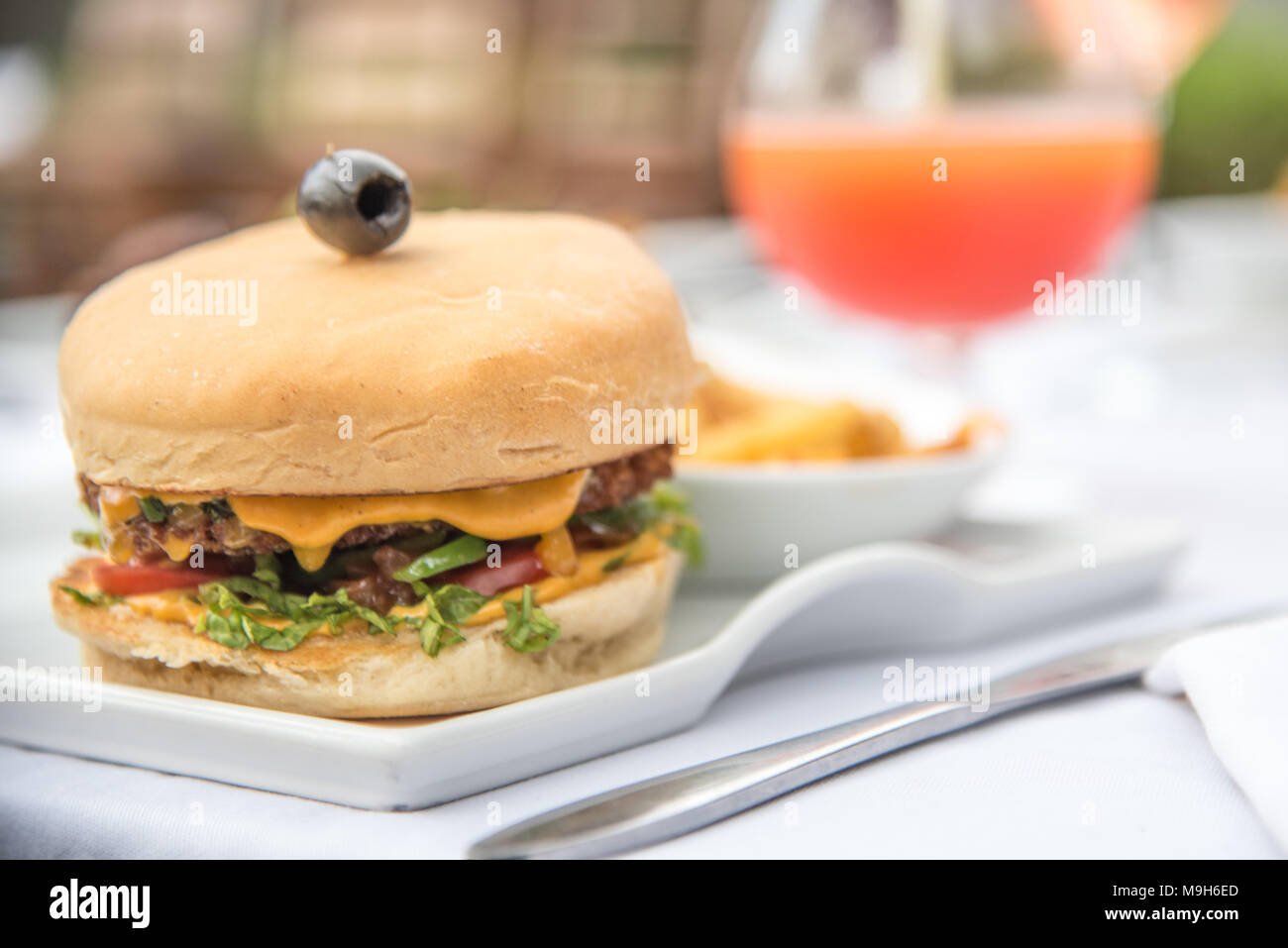 Un colpo di classic cheeseburger, guarnita con un nero oliva guarnire. Una luce arancione brillante cocktail/succo di frutta in background. Foto Stock