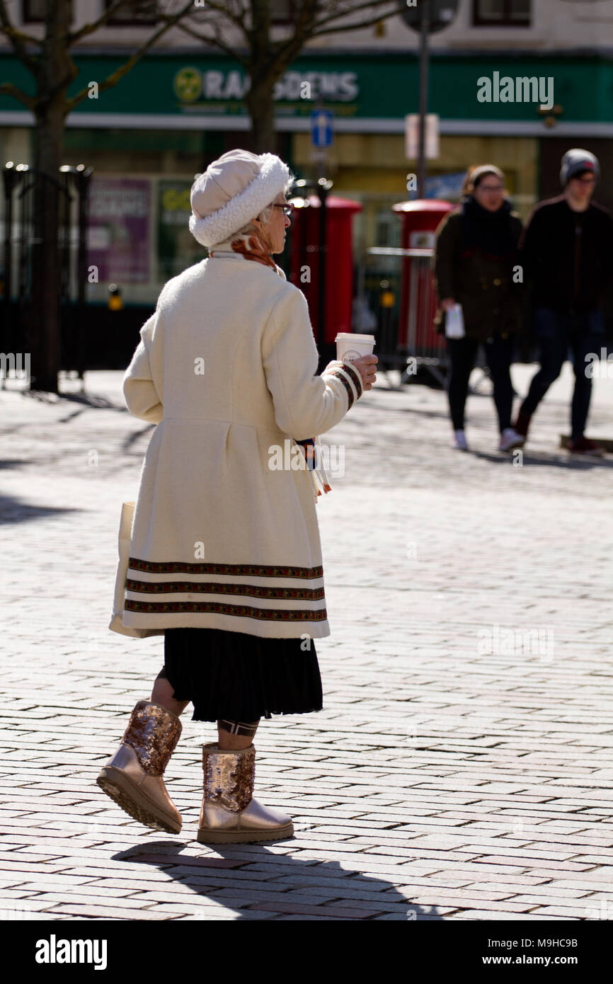 Una moda donna anziana tenendo una tazza di caffè da Clark's Bakery e indossando shiny ugg stivali a piedi circa nel centro città di Dundee, Regno Unito Foto Stock