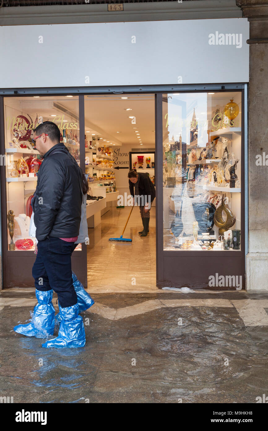 I turisti a piedi passato l'allagato in vetro di Murano shop su Piazza San Marco durante l'Acqua Alta, Venezia, Italia con una donna con straccio e acqua di riflessione Foto Stock