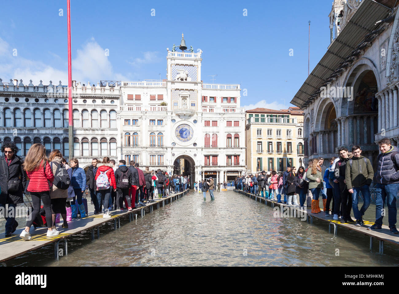 Due linee di persone su passarelles durante l'Acqua Alta (alta marea inondazioni), Piazza San Marco (Piazza San Marco), Venezia, Veneto, Italia con il clock T Foto Stock