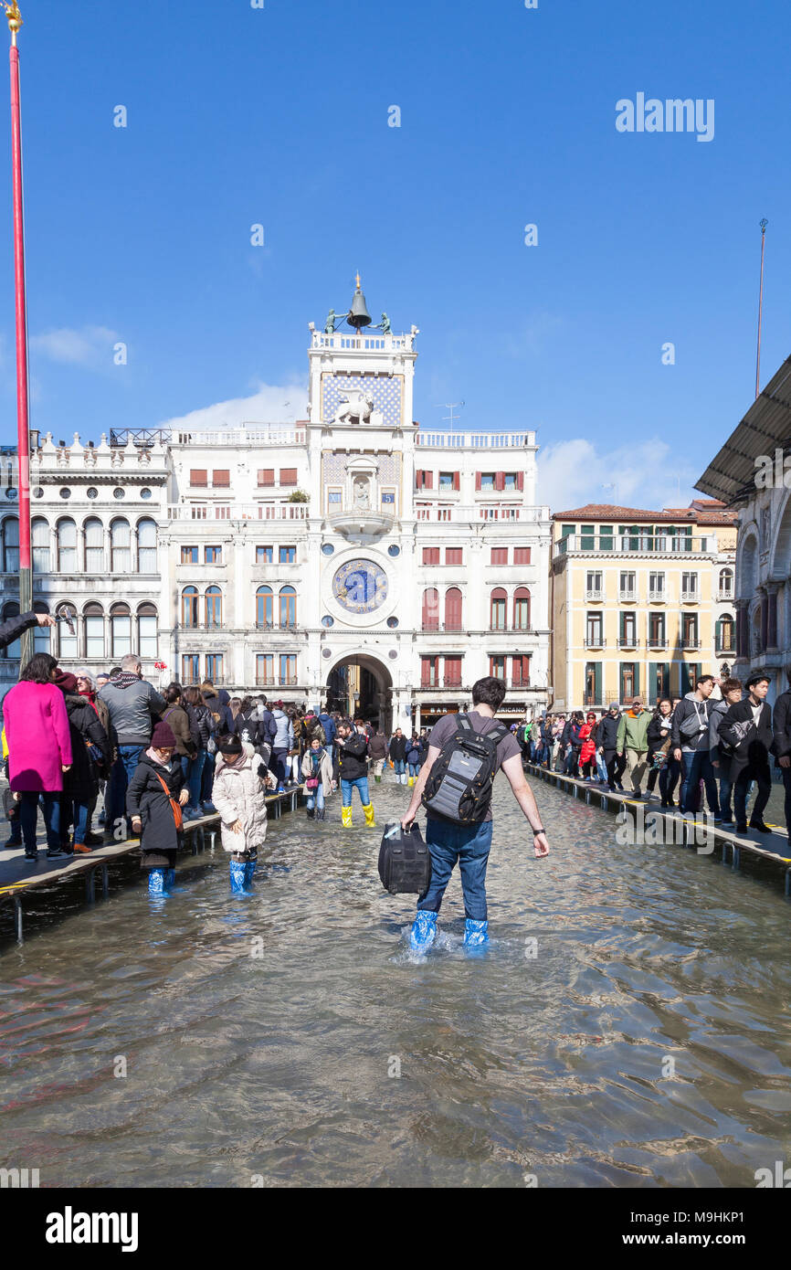 Turistico con bagagli guadare attraverso Acqua Alta inondazioni in Piazza San Marco (Piazza San Marco), Venezia, Italia con linee di persone su passerelles Foto Stock