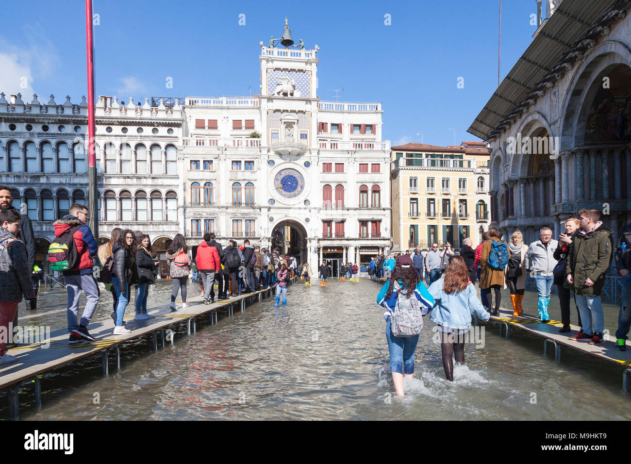 Due donne gli schizzi a piedi nudi attraverso Acqua alta a inondazioni, Piazza San Marco, Venezia, Italia guardato da persone che camminano su passarelles verso l'orologio T Foto Stock