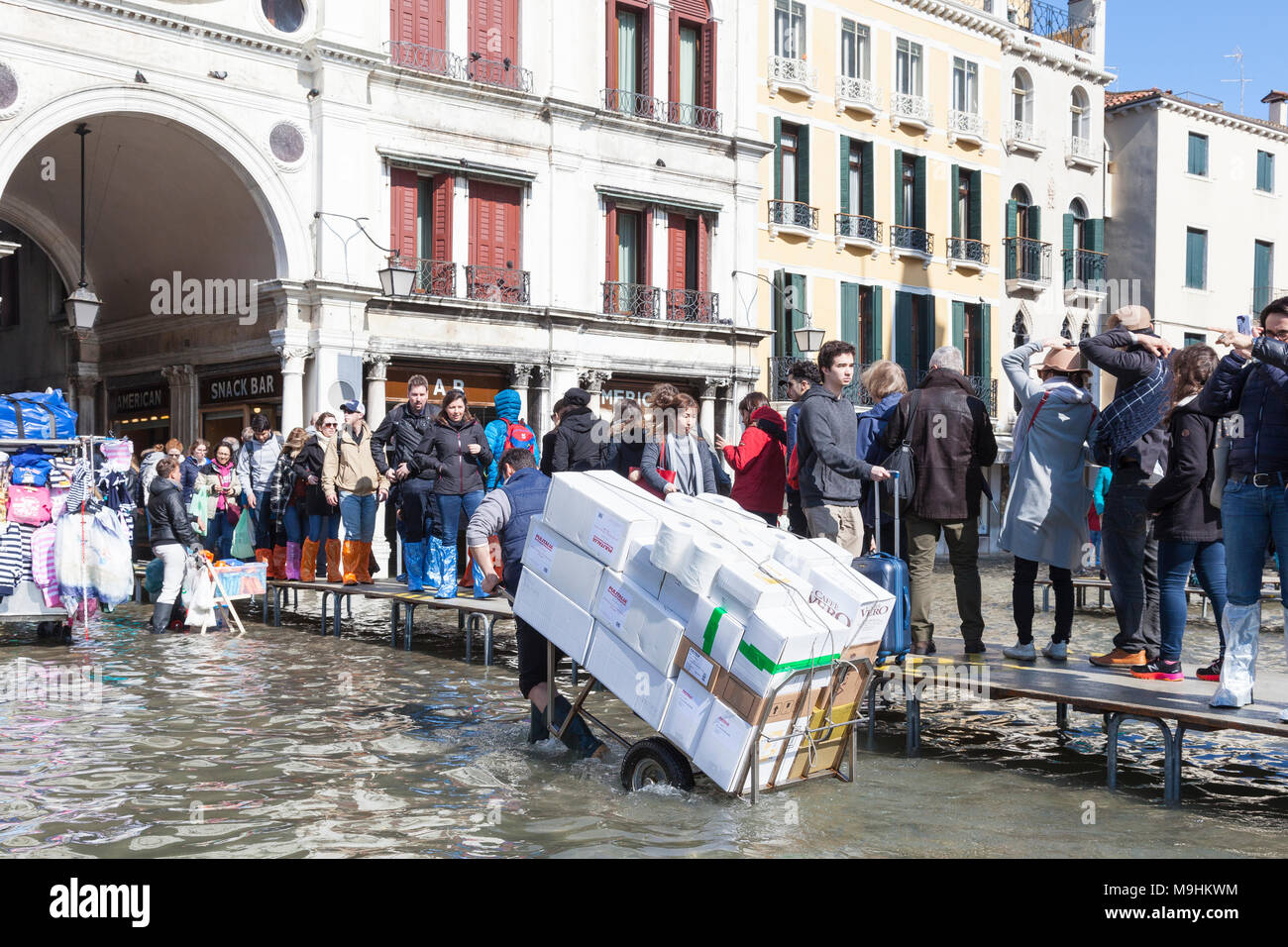 Lavoratore effettuare una consegna con un carrello in Acqua Alta inondazioni , Piazza San Marco (Piazza San Marco), Venezia, Italia passando la gente che camminava sul passarell Foto Stock