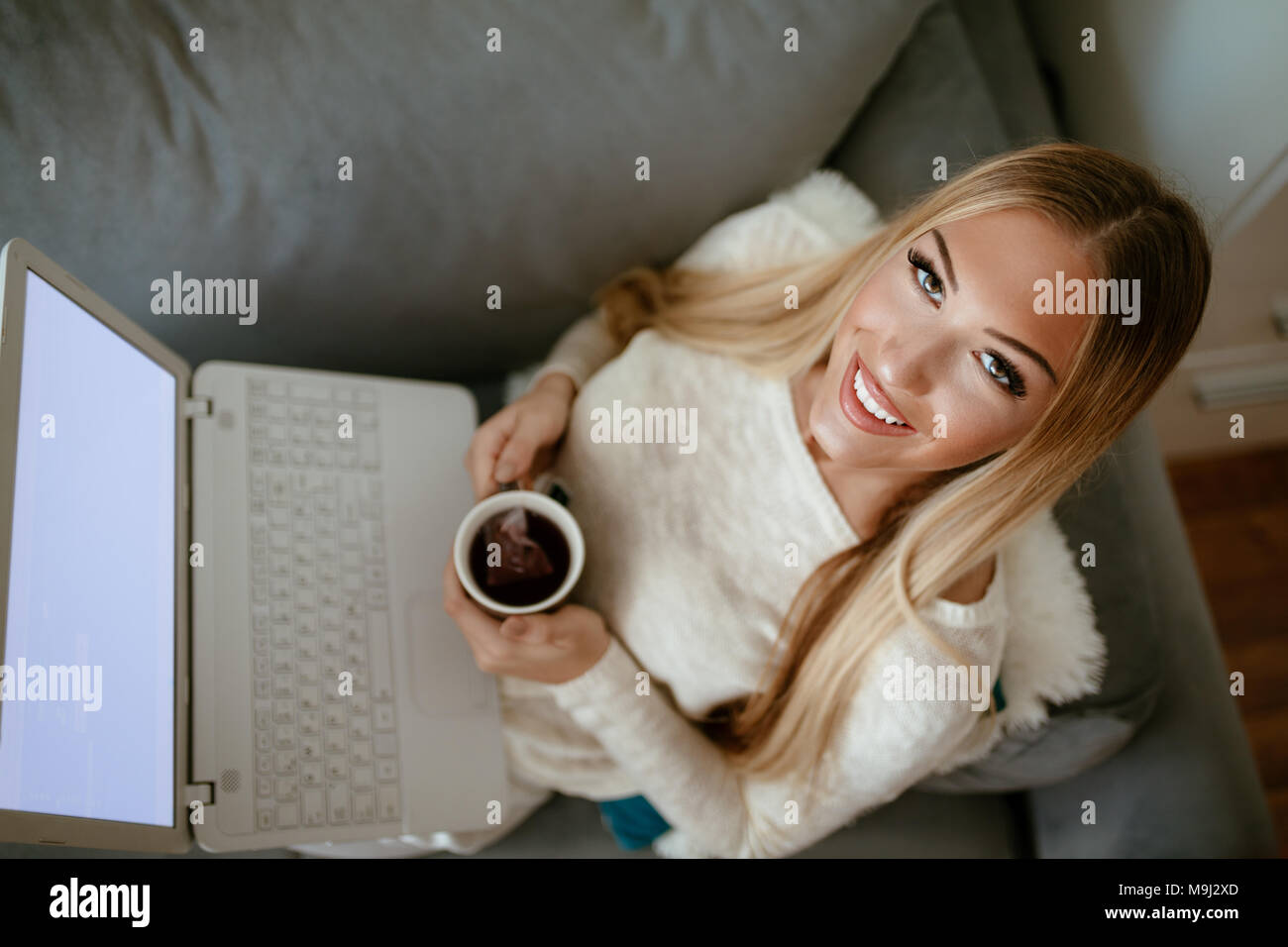 Attraente ragazza sorridente seduta alla sua stanza vivente con la tazza di tè utilizzando laptop. Ella è la navigazione in rete e del controllo dei mezzi di comunicazione sociale. Guardando camer Foto Stock