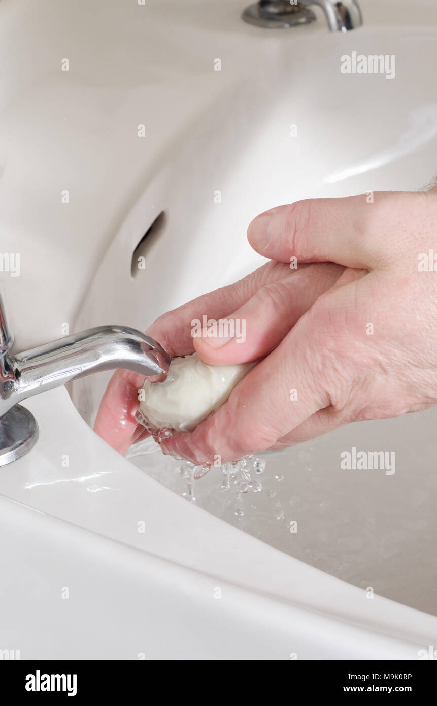 Mani maschili, tenendo una barra bianca del sapone e lavaggio chiaro sotto acqua corrente del rubinetto in porcellana lavello. Separare a caldo e a freddo di rubinetti. Foto Stock