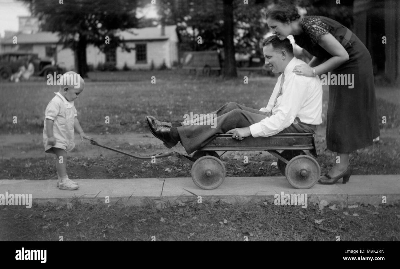 Carro-bound papà è bloccato tra un intraprendente moglie e tirando il toddler figlio, ca. 1920. Foto Stock
