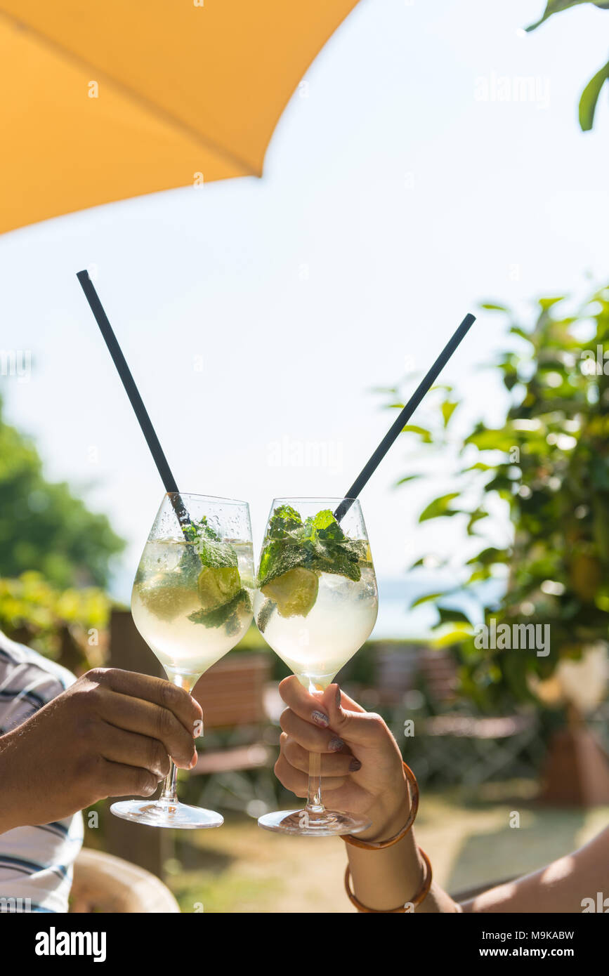 Giovane bere cocktail mojito in un accogliente bar all'aperto sulla spiaggia. Vecchia mano maschio e la mano di una giovane donna stanno tenendo i bicchieri sotto un ombrello. Foto Stock