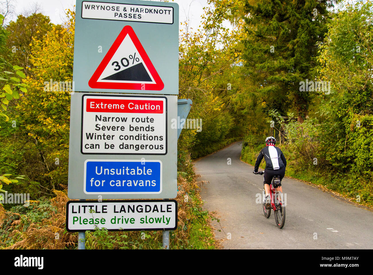 Ciclista passa i segnali di avvertimento sull'approccio di Wrynose e Hardknott passa in poco Langdale, nel distretto del Lago Foto Stock