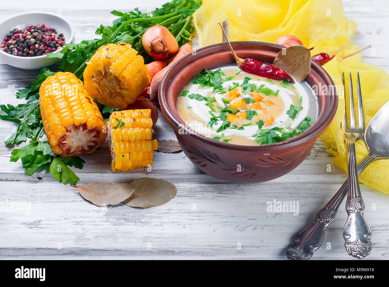 Zuppa dal mais con panna acida in una piastra di ceramica e gli ingredienti per la zuppa, pannocchie di granoturco, verdi, le carote. Mangiare sano Foto Stock