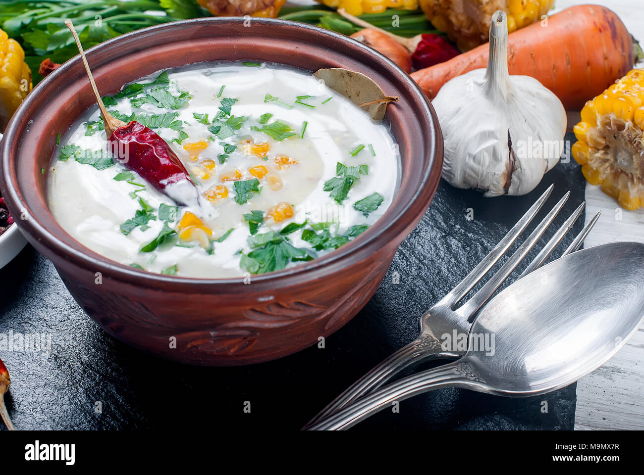 Zuppa dal mais con panna acida in una piastra di ceramica e gli ingredienti per la zuppa, pannocchie di granoturco, verdi, le carote. Mangiare sano Foto Stock