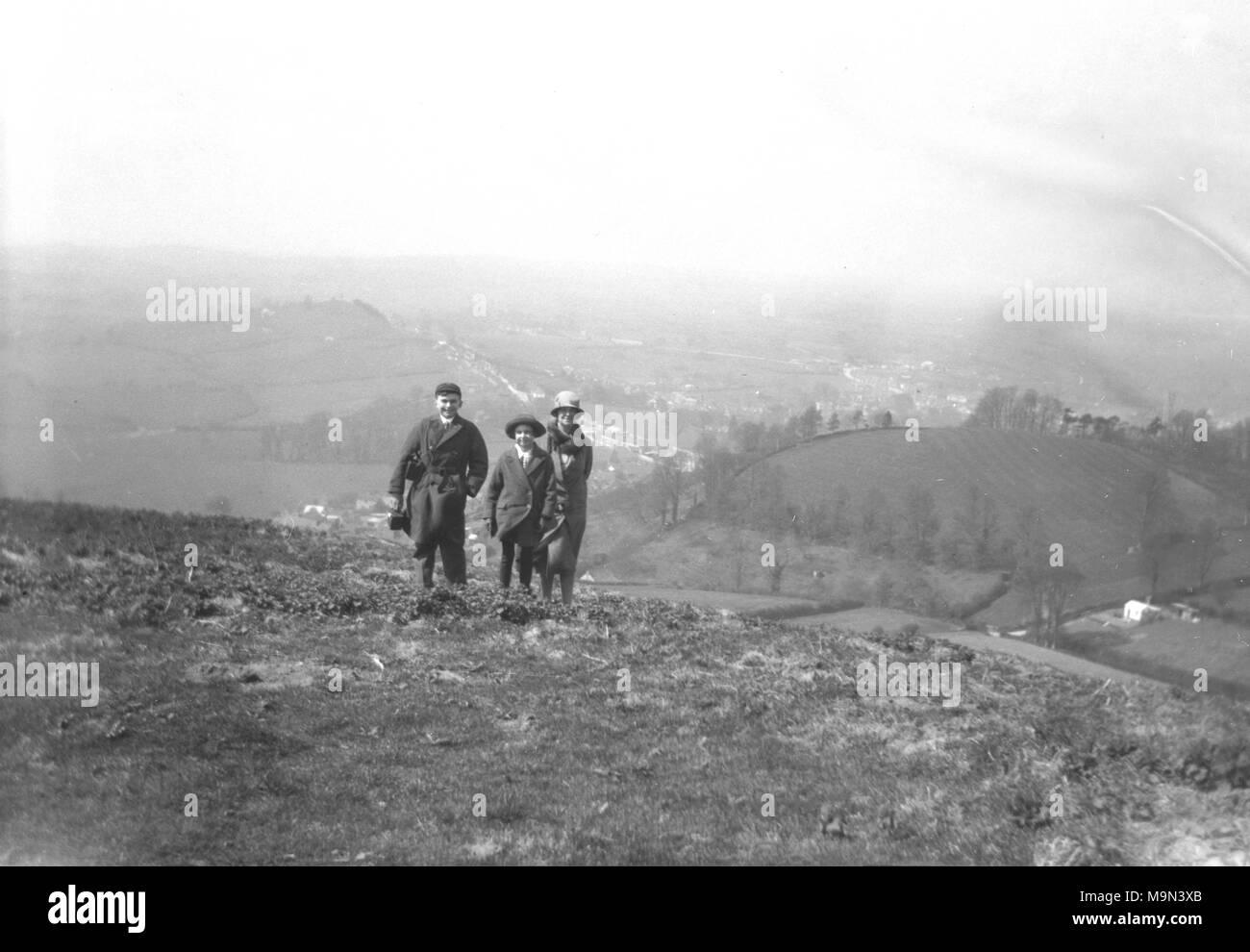 1920s, foto storiche di una madre con il figlio e la figlia in piedi sulla cima di una alta collina che domina la campagna circostante. La signora e la figlia di indossare abbigliamento normale, mentre il ragazzo è in uniforme scolastica che indossa un cappello e portando la sua scatola di pranzo, Inghilterra, Regno Unito. Foto Stock