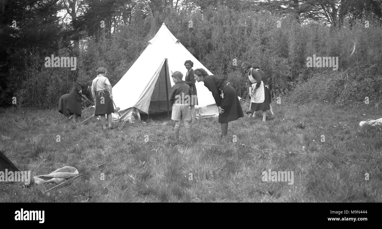 1930s, storico girl guides mettendo su una tela di canapa tenda al di fuori di un campo erboso, Inghilterra, Regno Unito. Foto Stock